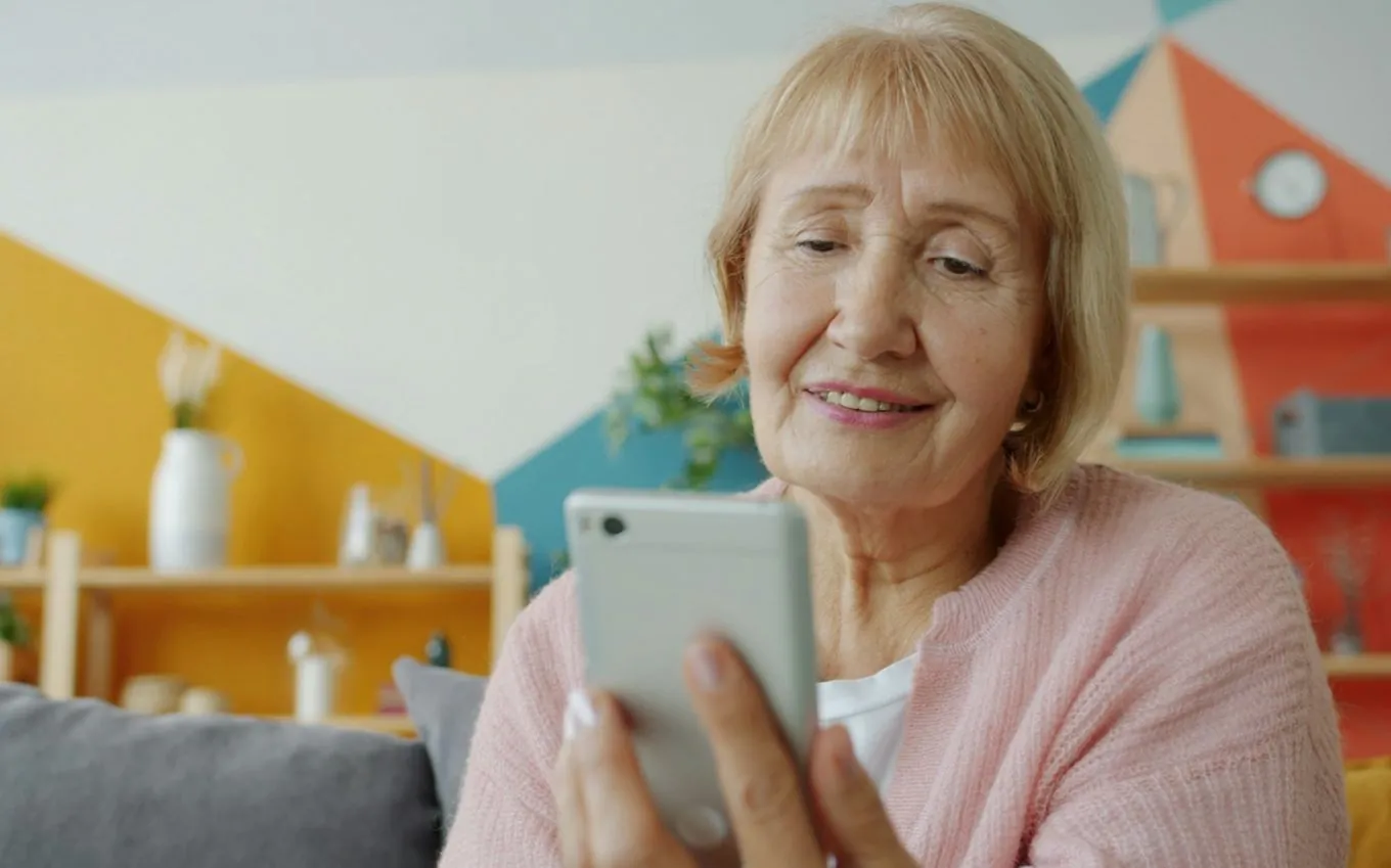 An older woman smiling at her smartphone on a sofa