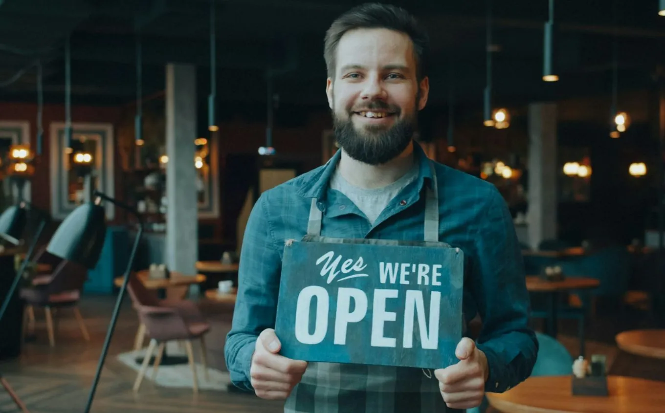 A smiling café owner in an apron holding a sign that says Yes we're open